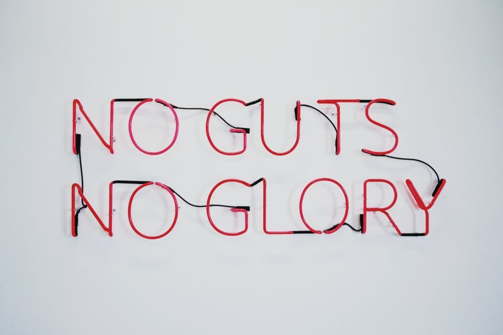 Vegan neon sign with the words "NO GUTS, NO GLORY" in red and pink neon tubing on a white wall.